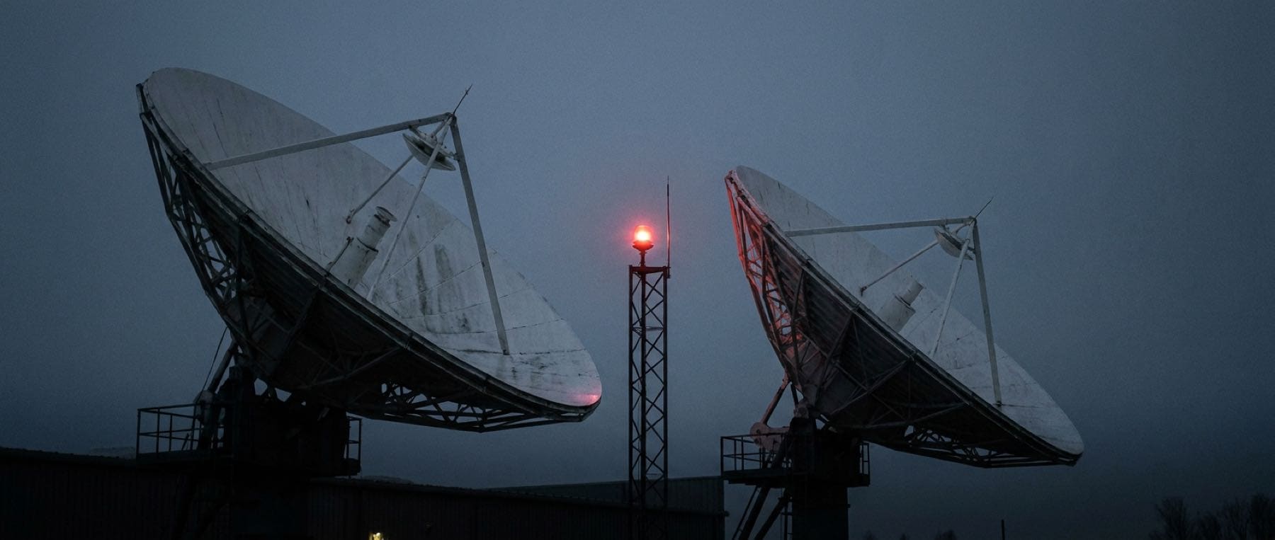 Two satellite uplink dishes at dusk, pointed high against a deep blue-grey sky